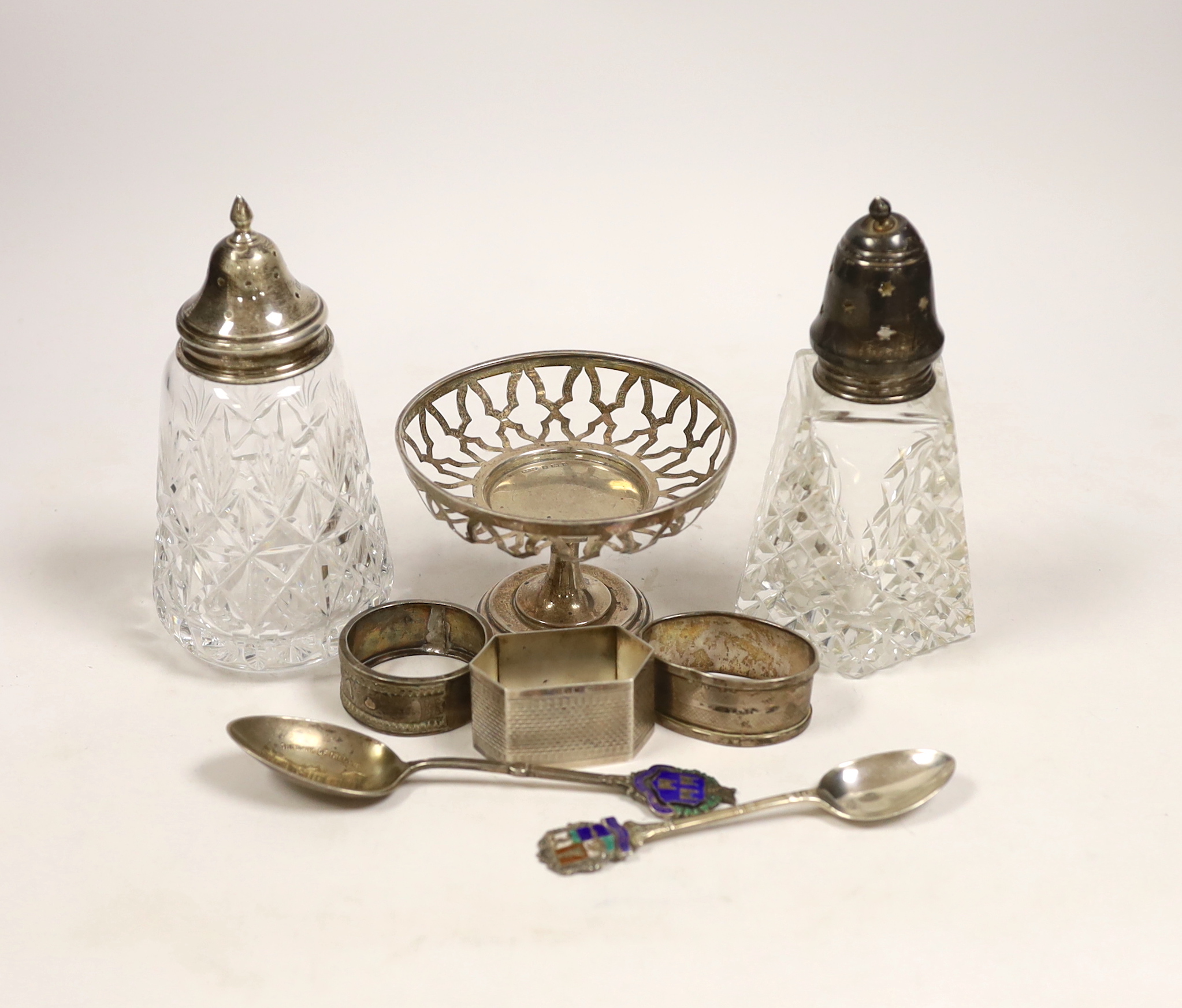 A group of small silver including two mounted glass sugar sifters, three assorted napkin rings, two commemorative spoons and a small pedestal nut dish (lacking insert).